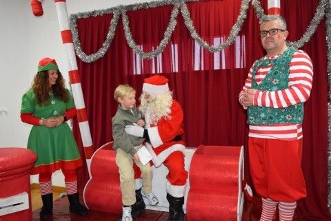 Papá Noel hablando con un niño en su rodilla acompañado de sus elfos