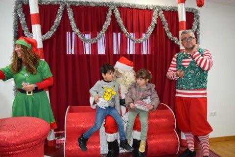 Papá Noel tomándose una foto con un niño y una niña en sus rodillas acompañado de sus elfos